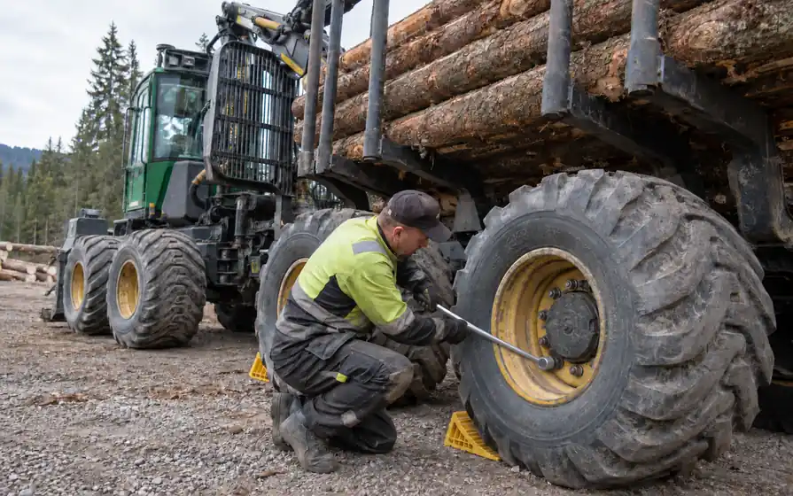 Mechanic tightening a lower peripheral wheel nut on a forestry forwarder tyre beside its loaded log bunk.