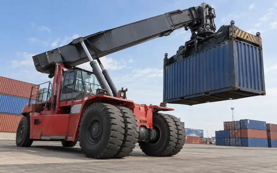 Reach stacker with a telescopic boom and prominent paired front tyres in a container terminal.
