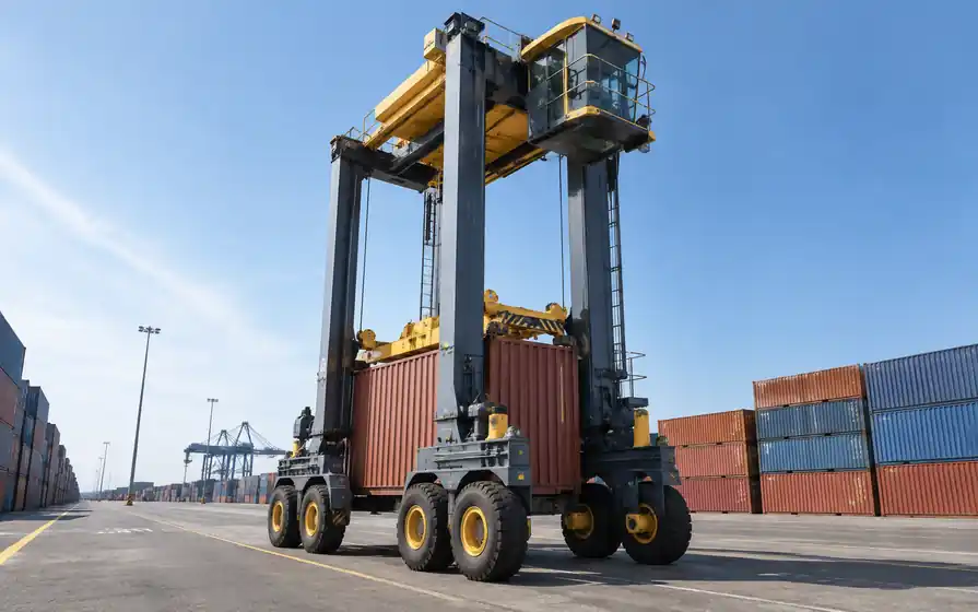Tall straddle carrier astride a shipping container, with rubber wheel sets along its narrow portal frame.